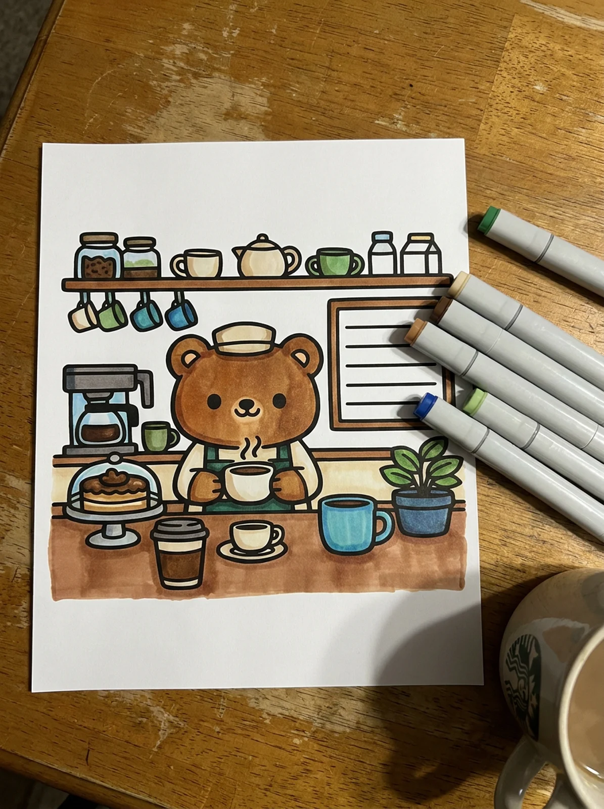 A printed bear barista coloring page, partly colored with markers, on a wooden table next to a mug and uncapped markers