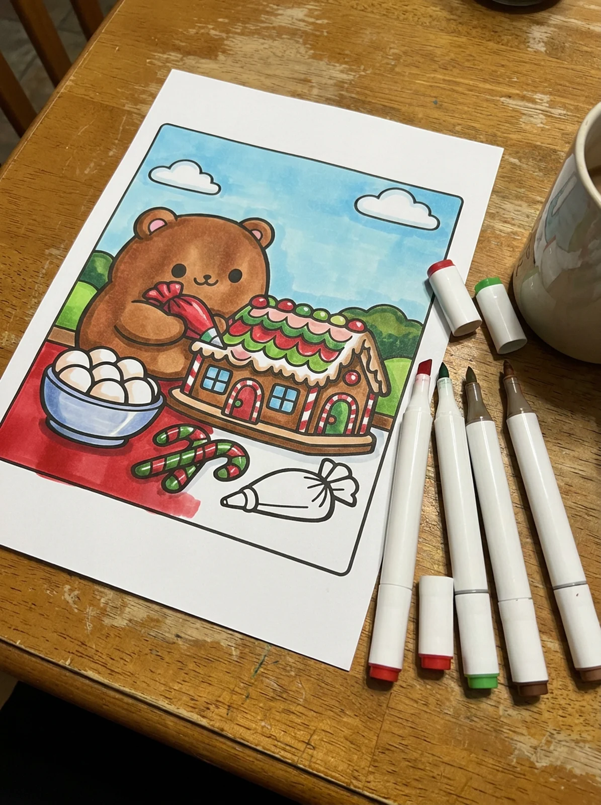 A printed coloring page of a bear decorating a gingerbread house, partly colored with markers, on a table